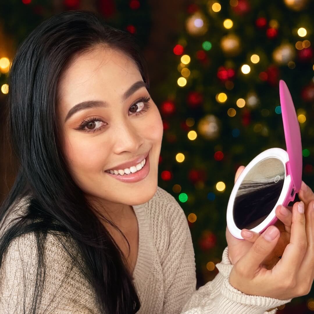 Woman smiling, holding a pink compact mirror with festive lights blurred in the background.
