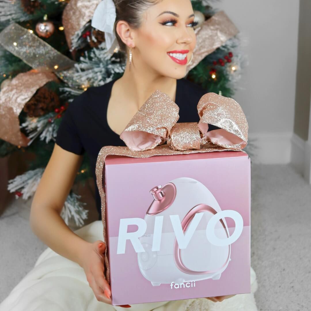 Person holding a pink gift box with a large bow, sitting by a decorated Christmas tree.