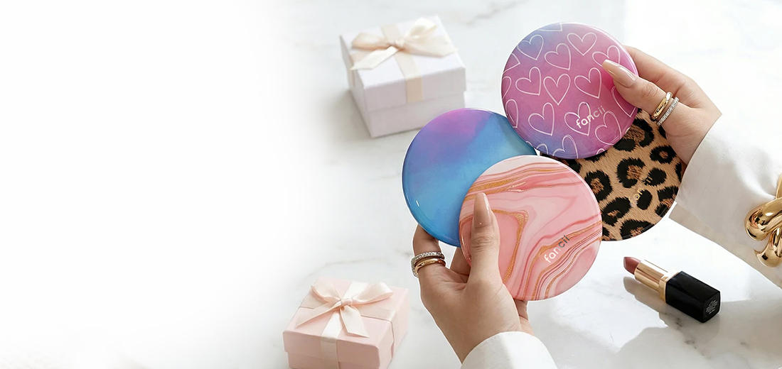 A person's hands hold four compact mirrors with different designs, with gift boxes and lipstick nearby.