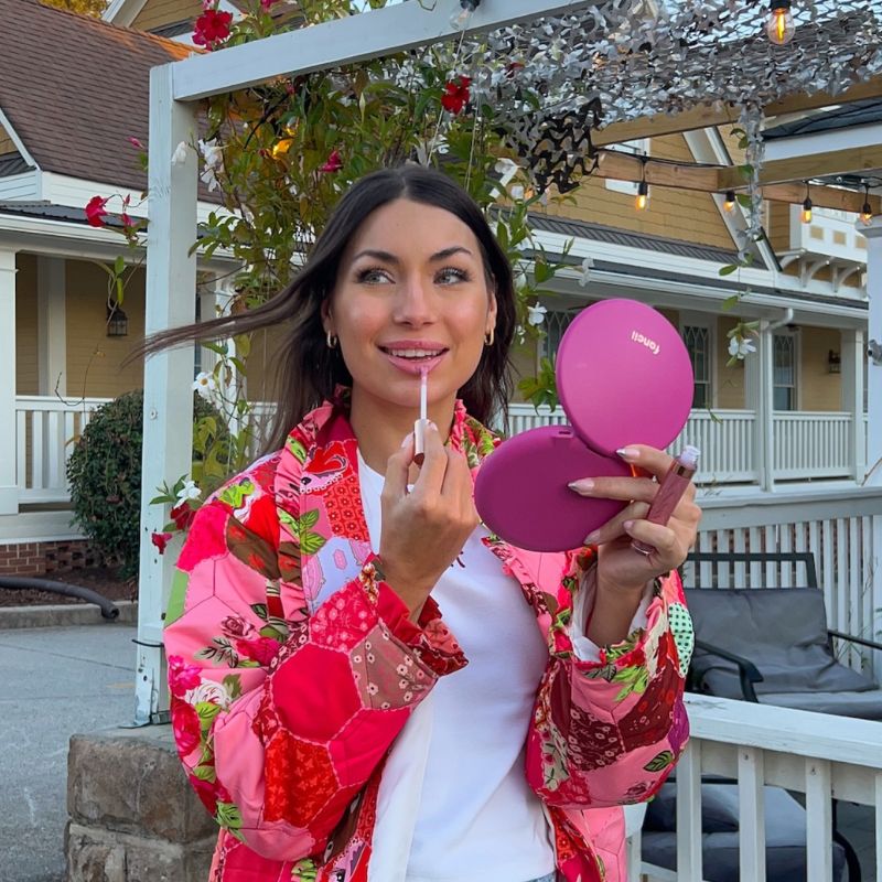 Person applying lip gloss with pink compact outdoors.