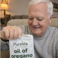An older man with white hair smiles while holding a pouch of Purevia Nutrition oil of oregano.