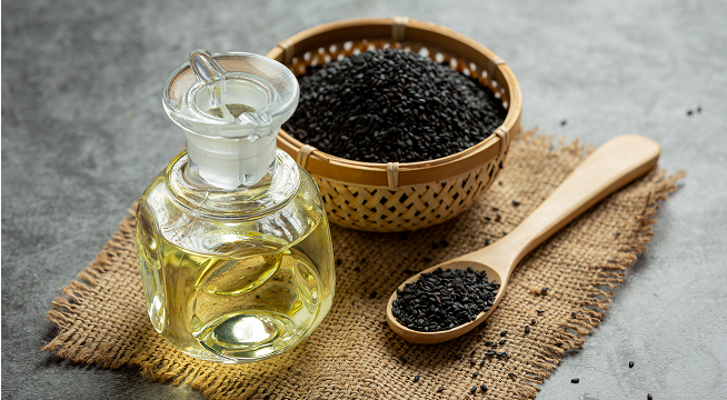 Glass jar of oil, basket of black seeds, and wooden spoon on burlap fabric.