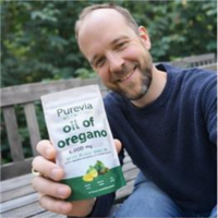 A smiling man sits on a bench outdoors, holding up a pouch of Purevia oil of oregano.