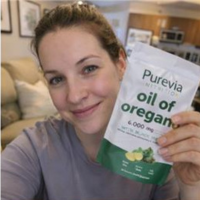 A woman smiles while holding up a white and green pouch of Purevia Nutrition oil of oregano.