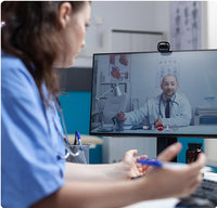 A medical professional in scrubs has a video conference with a doctor on a computer monitor.