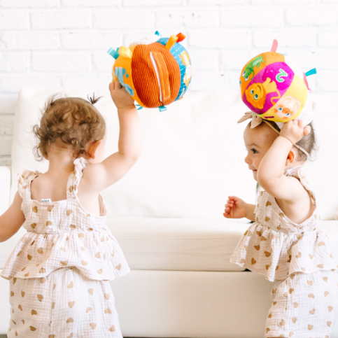 Two toddlers in matching outfits holding colorful balls indoors.