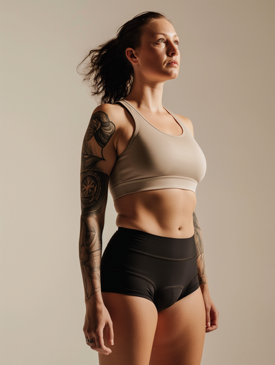 A woman with tattoos on her arms, wearing a sports bra and shorts, stands against a plain background.