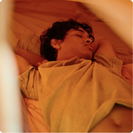 A young man with dark curly hair sleeping on his back with his hands behind his head.