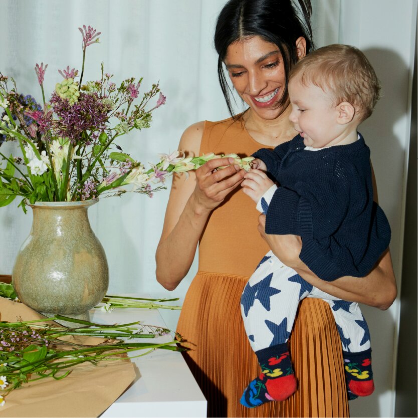 A smiling woman holds a baby while they look at a flower she is arranging in a vase.
