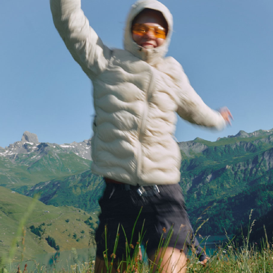 A person in a light-colored puffer jacket and sunglasses jumps with a mountain range in the background.