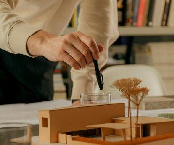 A person's hand holding a pen over a cardboard architectural model of a modern house.