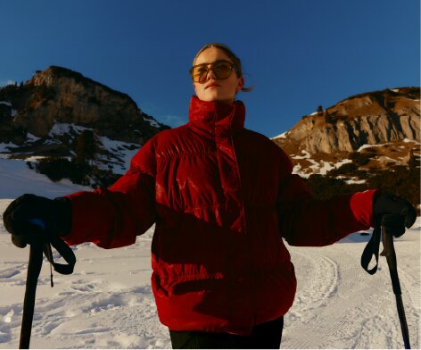 A person in a red puffer jacket and sunglasses holds ski poles in a snowy mountain landscape.