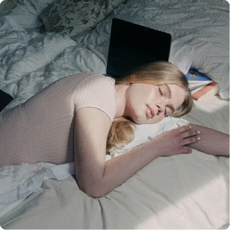 A young woman sleeps in a sunlit bed with a laptop and books next to her.