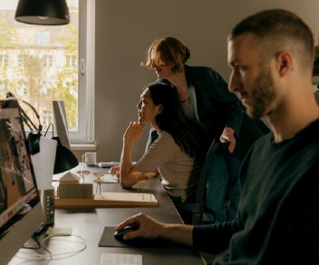Three people working at desks with computers in an office, with natural light from a window.