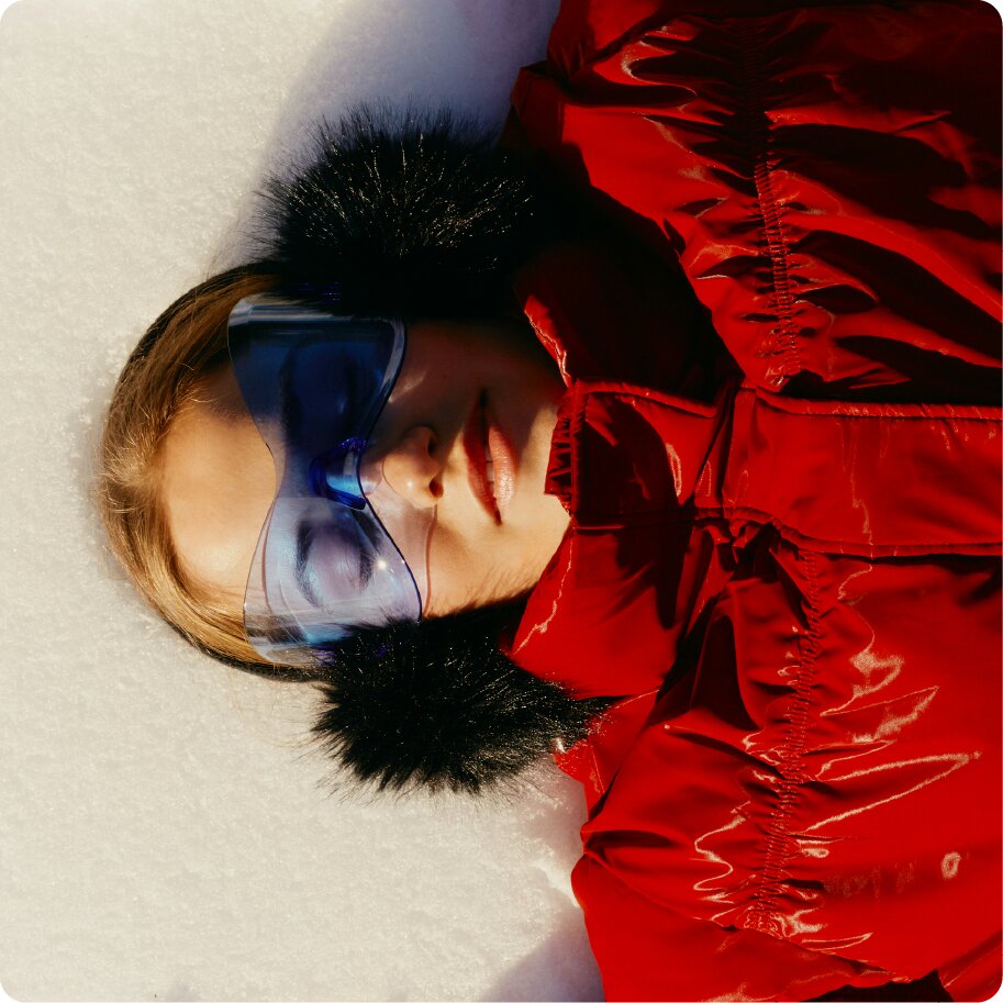 An overhead shot of a person in a red jacket and blue ski goggles lying in the snow.