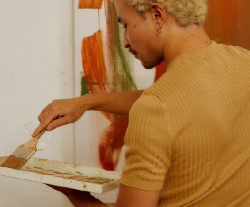 A person with blonde hair wearing a tan shirt paints on a canvas with a brush and palette.