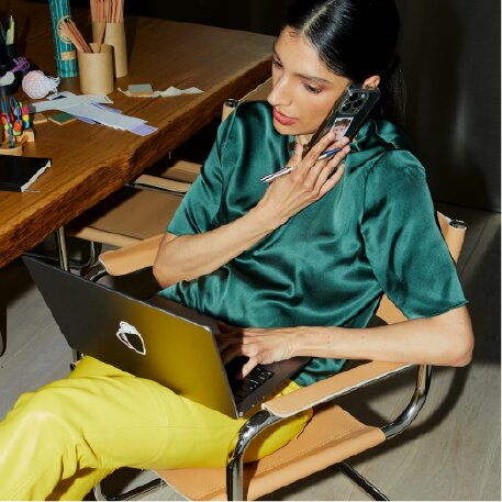 A person in a green shirt and yellow pants talks on a phone while using a laptop at a desk.
