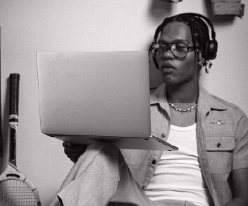 A black and white photo of a person with headphones and glasses looking at a laptop screen.