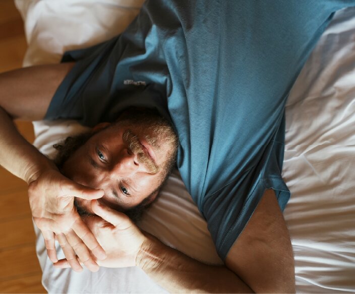 An overhead shot of a man with a beard lying in bed, looking up at the camera.