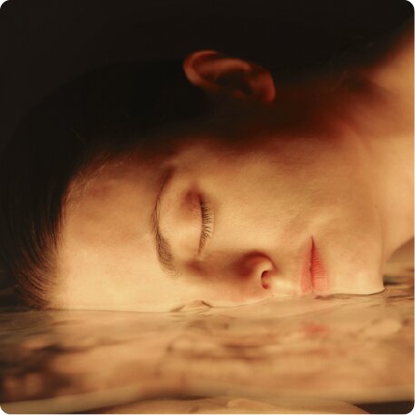 A close-up of a person's face with eyes closed, resting on a watery, reflective surface.