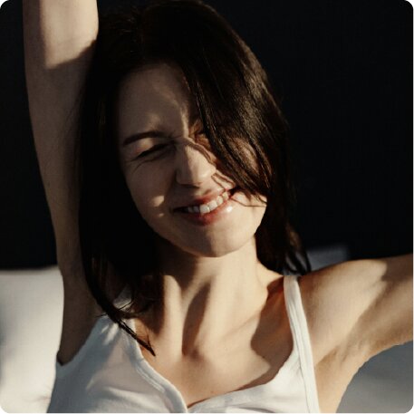 A young woman with dark hair in a white tank top stretches and smiles, her eyes squinted shut.