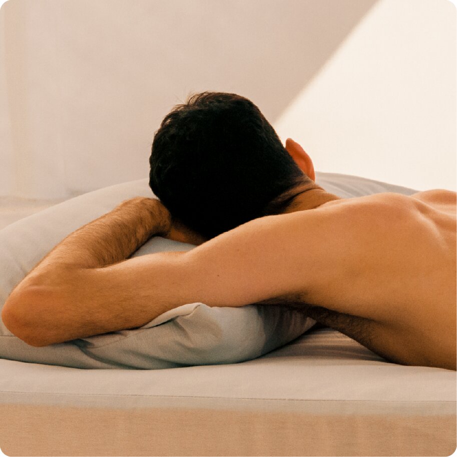 The back of a shirtless man with dark hair lying face down on a pillow in bed.