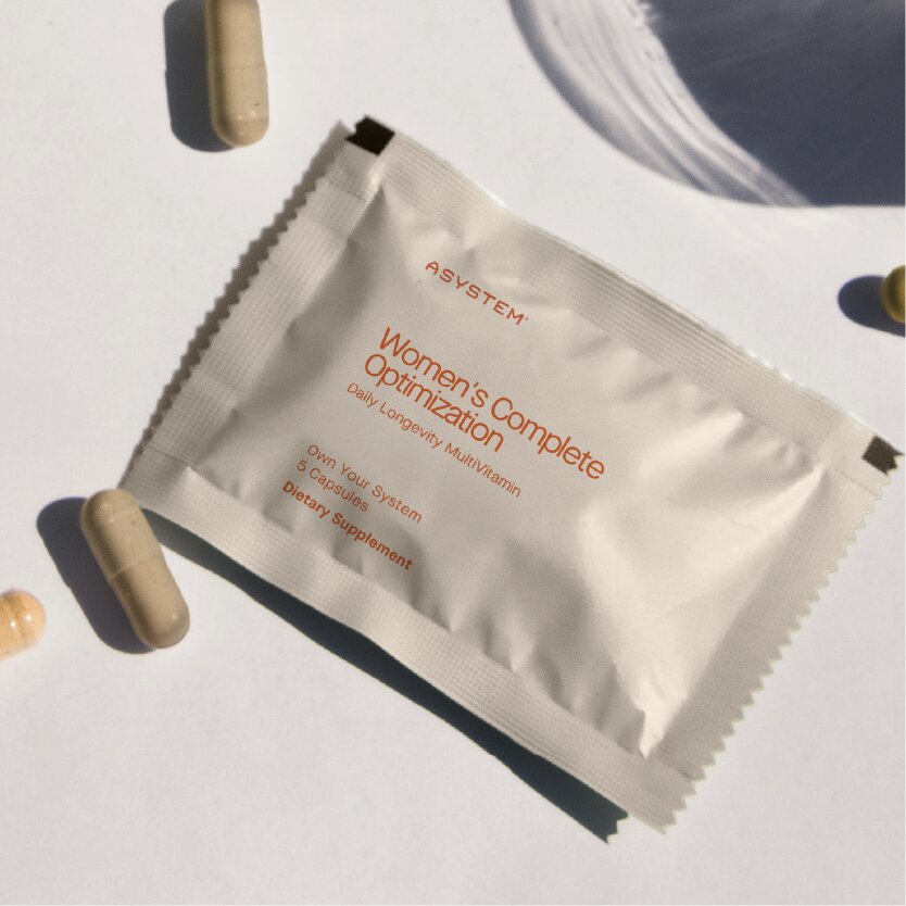 A single-serving packet of ASYSTEM Women's Complete Optimization dietary supplement, with several capsules scattered around it.