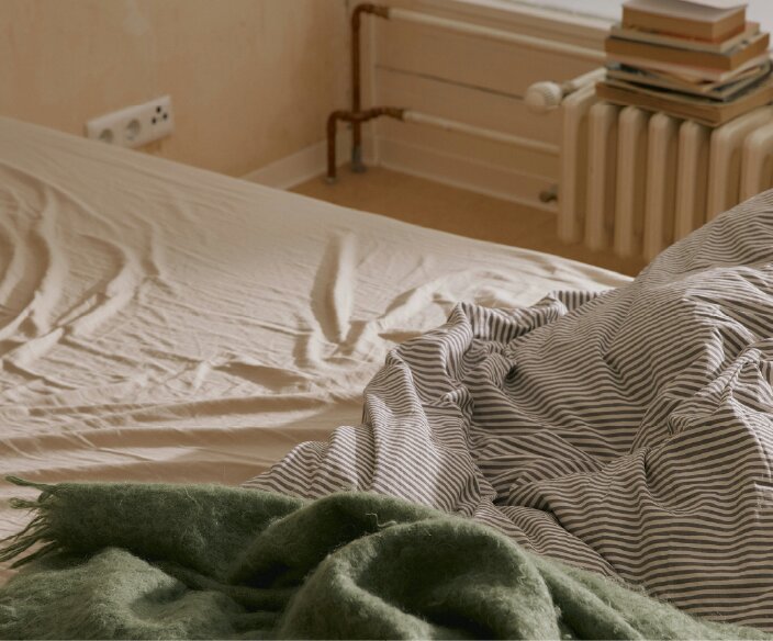 A close-up of an unmade bed with a striped duvet, green throw, and a radiator with books.