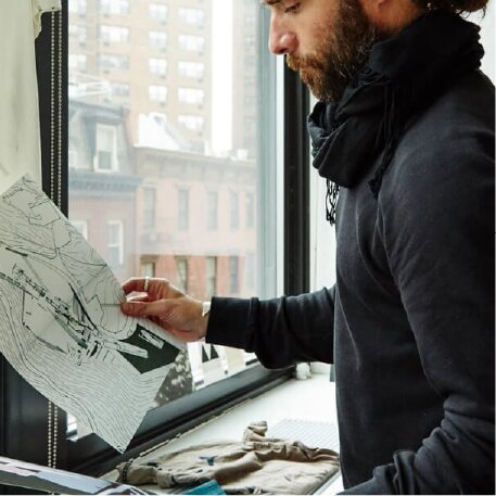 A bearded man wearing a dark sweatshirt and scarf looks at a drawing next to a window.