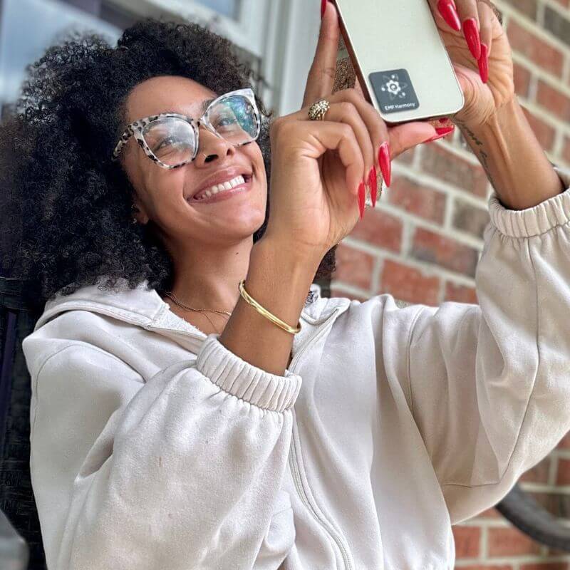 Woman smiling and taking a selfie with a smartphone, wearing glasses and a white hoodie.
