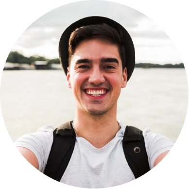 Person smiling outdoors, wearing a hat and backpack, with water in the background.