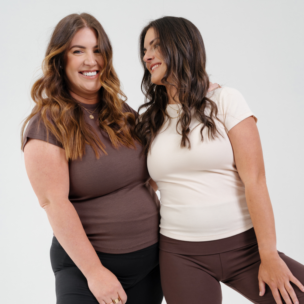 Two women smiling, wearing casual tops and leggings, standing together against a plain background.
