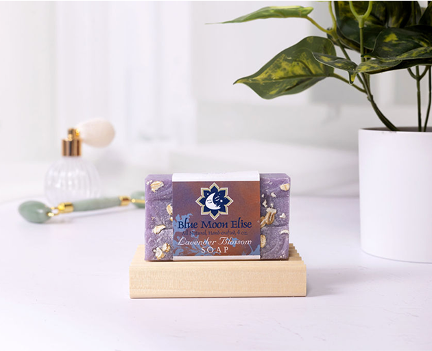 Purple soap bar on a wooden dish with a plant and perfume bottle in the background.