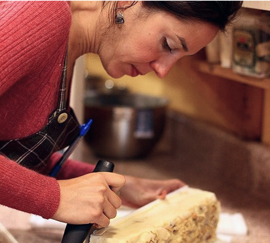 Person in a kitchen slicing a large block of cheese on a counter.