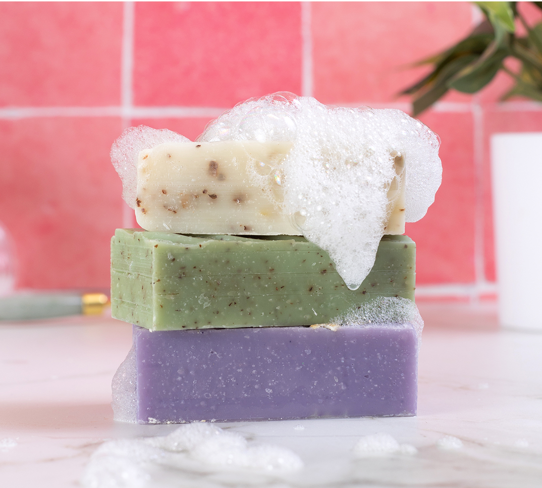 Three stacked soap bars with bubbles, against a pink tiled background.