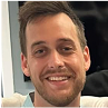 A person smiling, with short hair and a beard, wearing a light blue shirt.