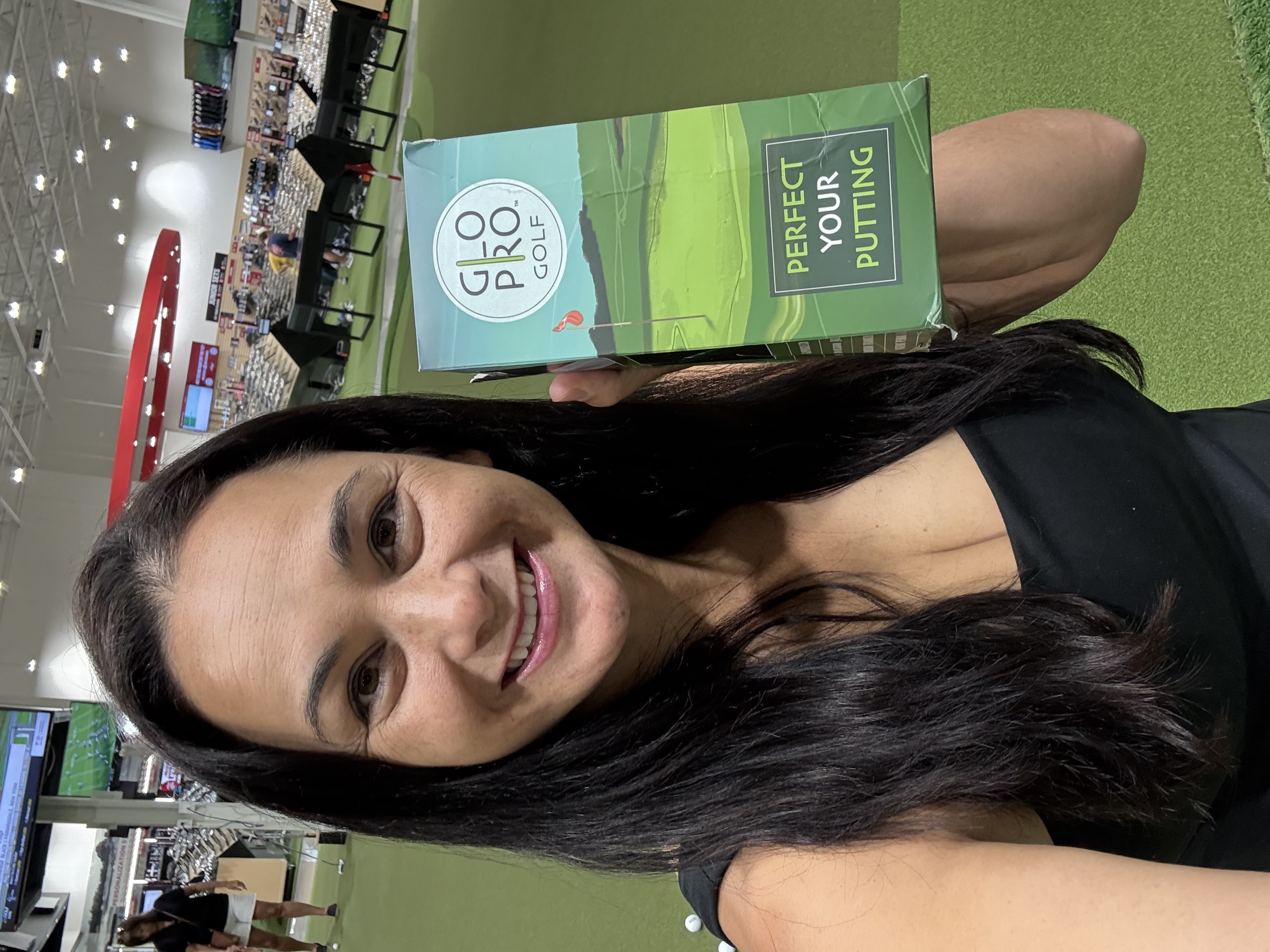 Person holding a golf product box, standing on a putting green indoors.