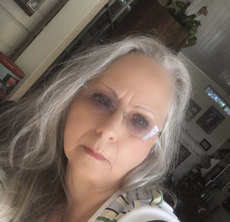 A woman with long gray hair and glasses in a room with various decorations.