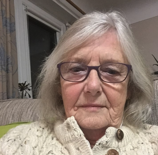 Elderly woman with glasses and white hair sitting indoors.
