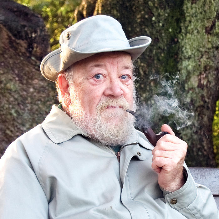 Elderly man with a pipe and hat, sitting outdoors.