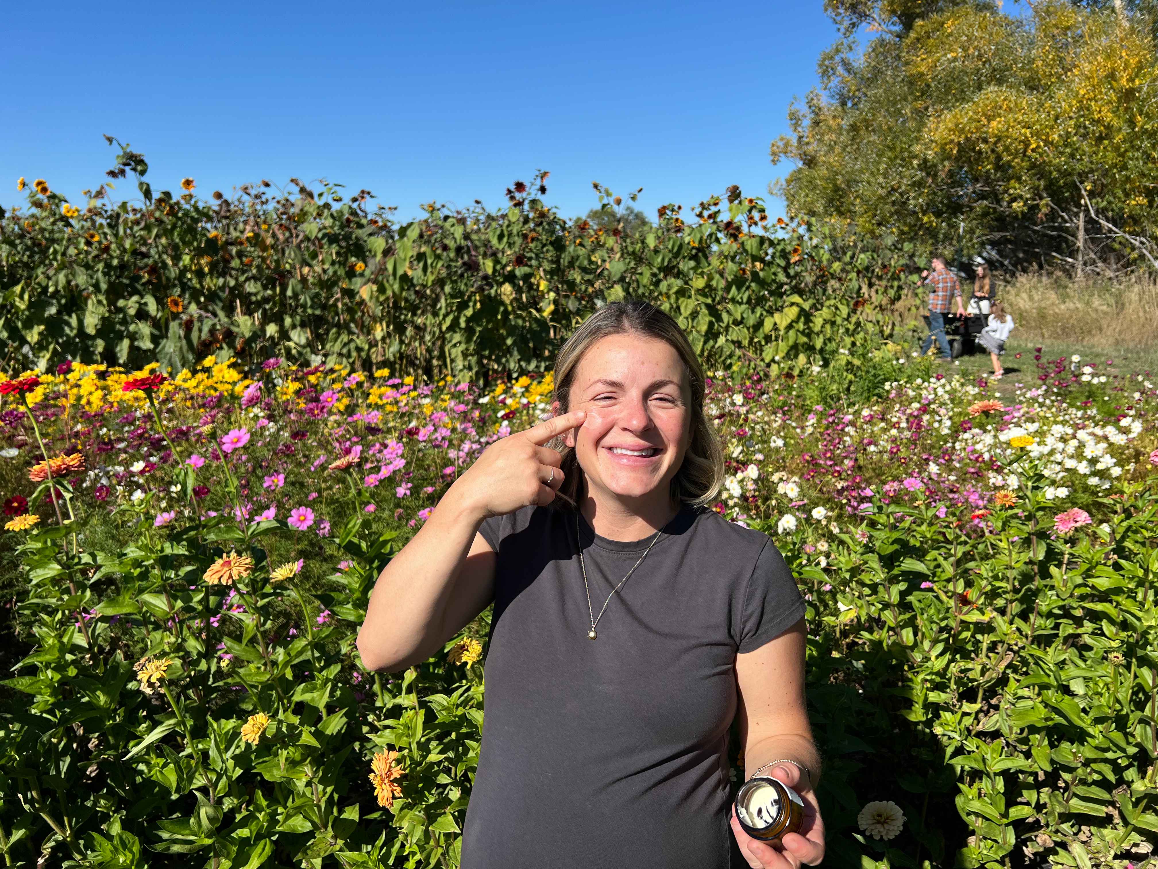 A smiling woman in a field of colorful flowers points to her face while holding a small jar.