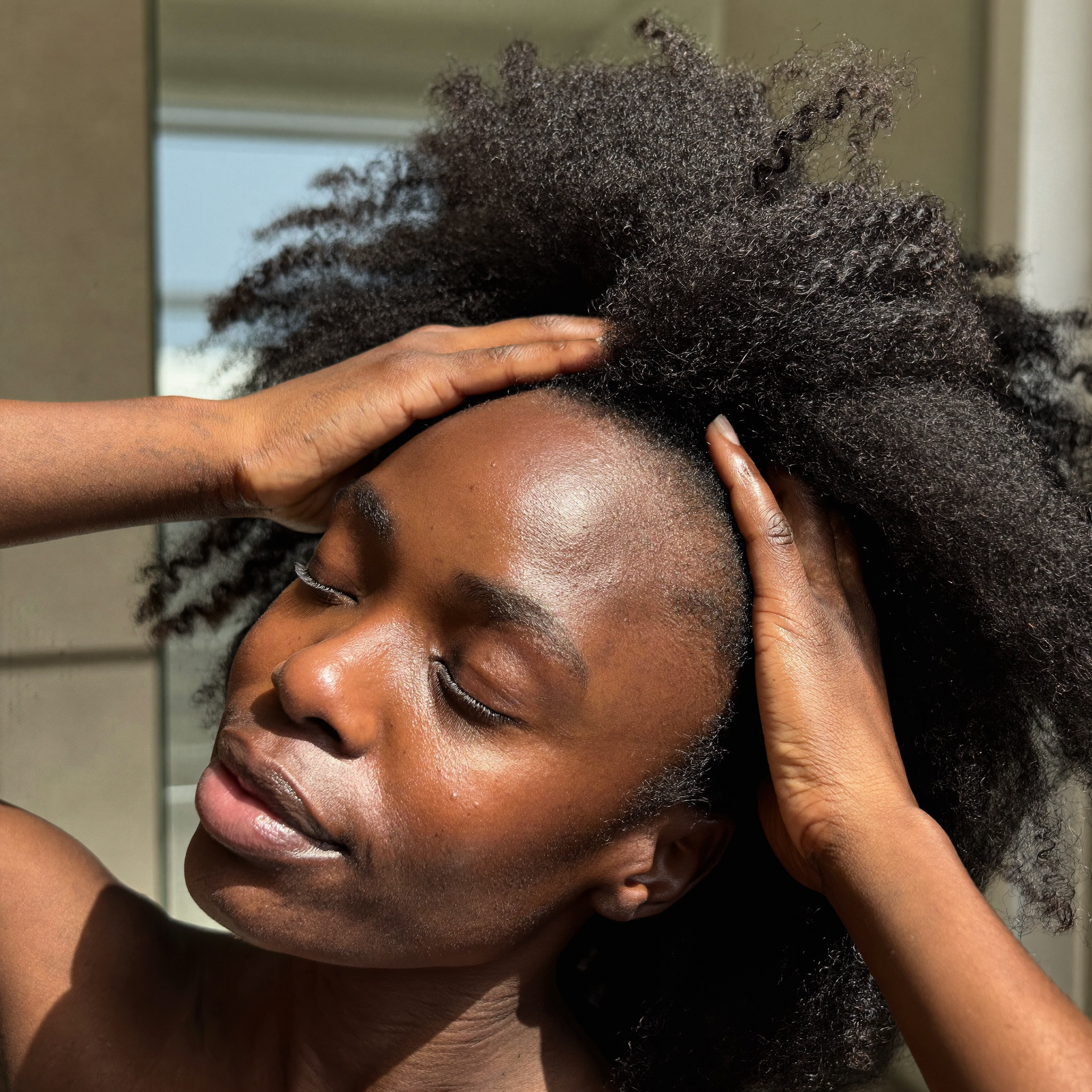 A Black person with eyes closed runs their hands through their thick, coily hair in the sunlight.