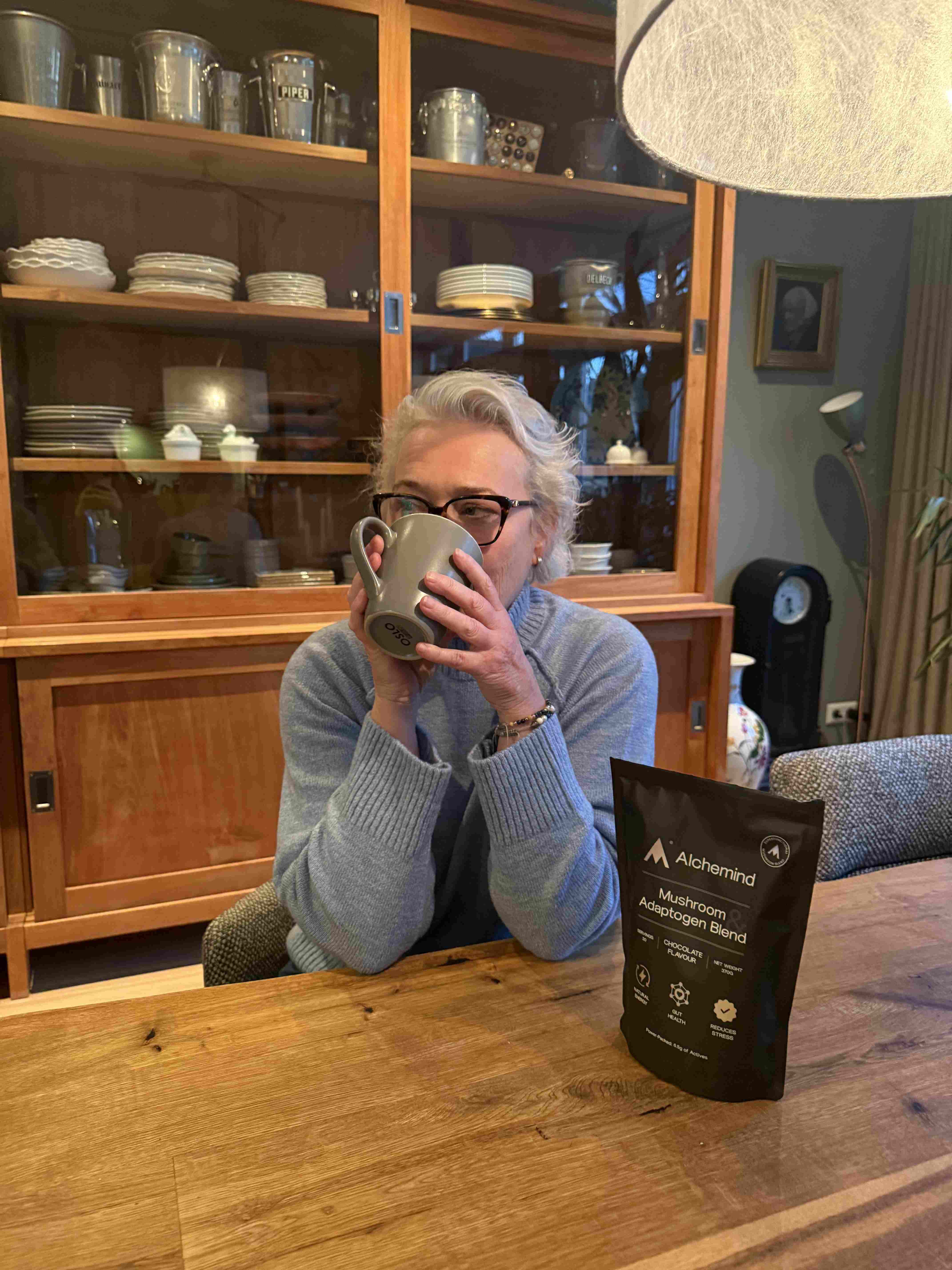 Person in blue sweater drinking from a mug near a wooden cabinet and a product package.