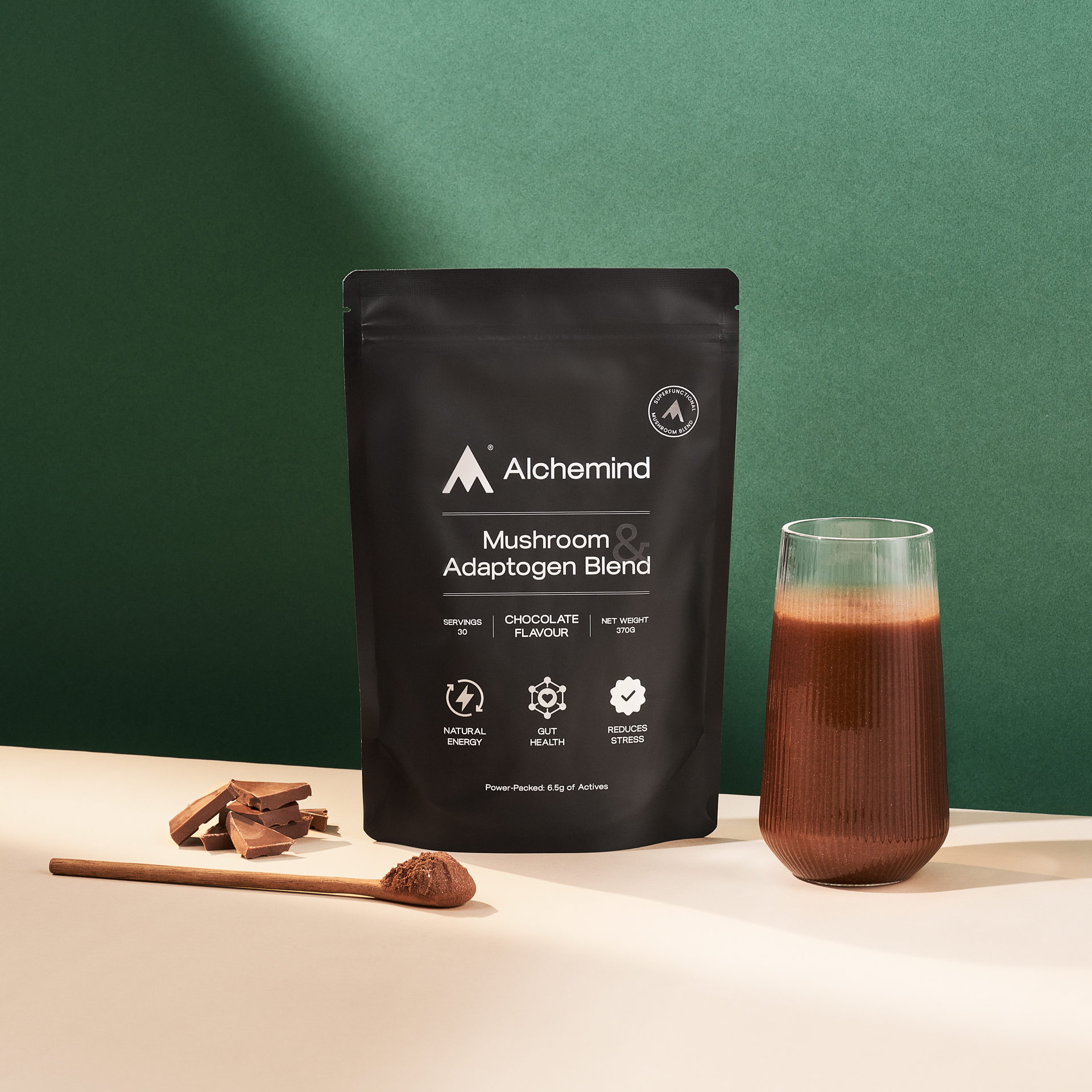 Black pouch of mushroom blend, glass of chocolate drink, spoon, and chocolate pieces on a table.