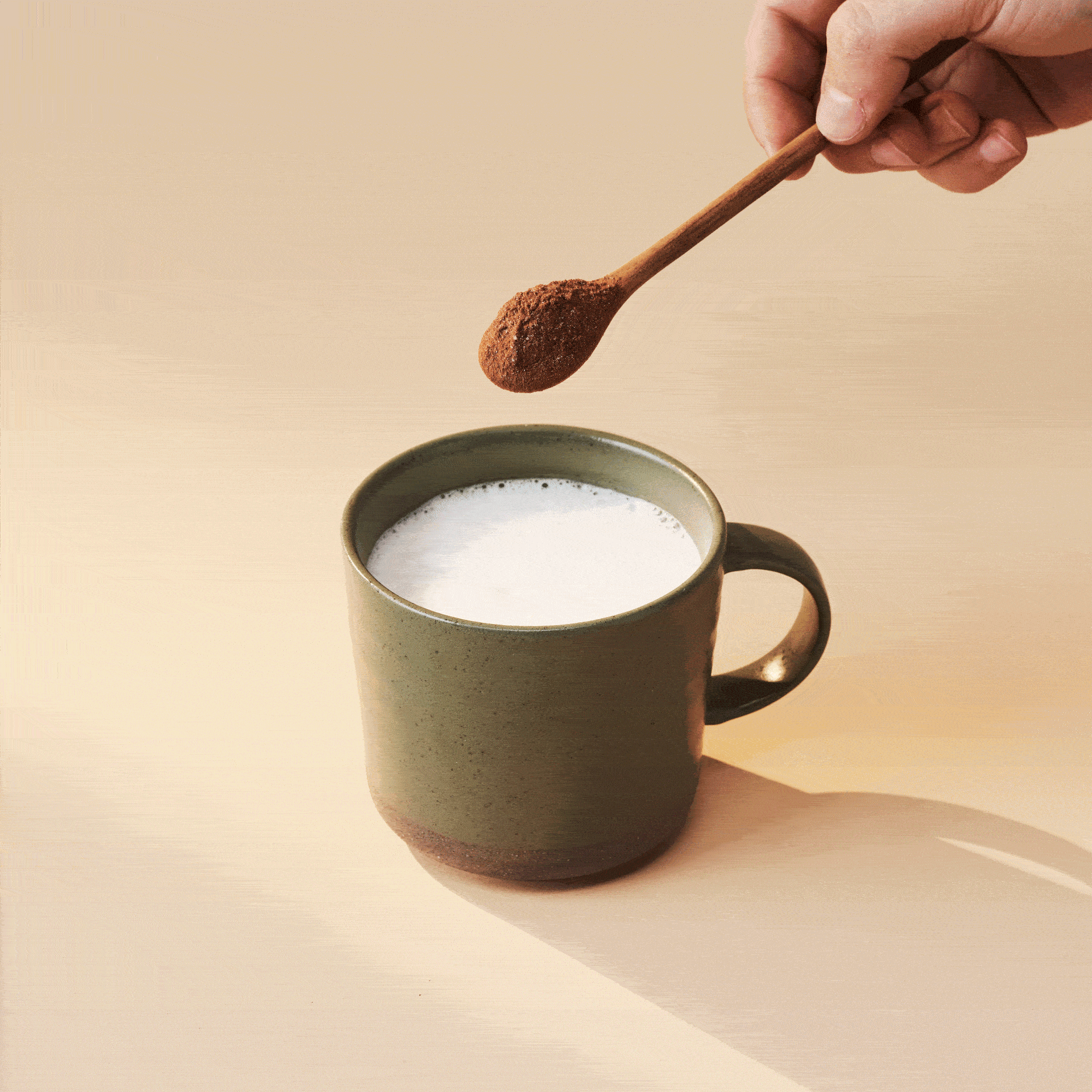 A hand holds a spoon with cocoa powder above a green mug of milk.