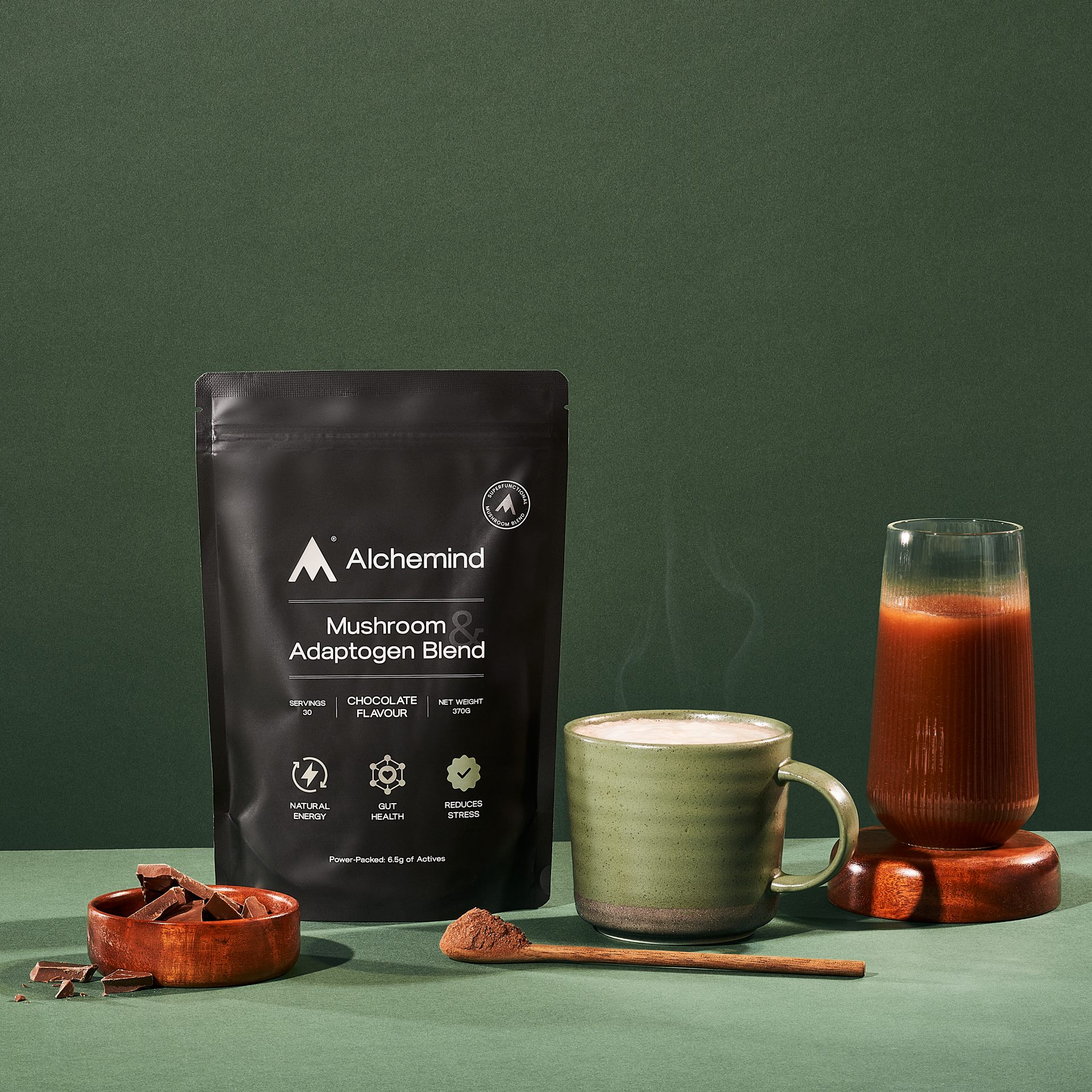 Pouch of mushroom adaptogen blend with a green mug and glass of brown liquid on a green surface.