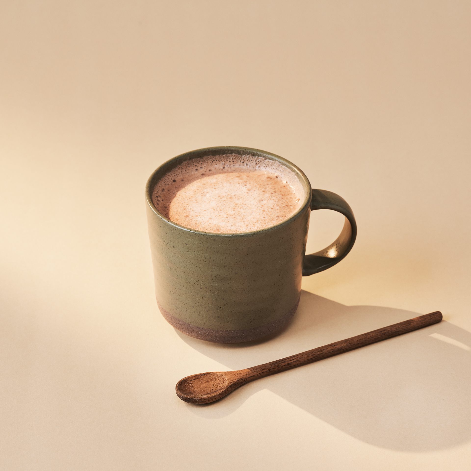 Green mug of frothy coffee with a wooden spoon on a beige background.