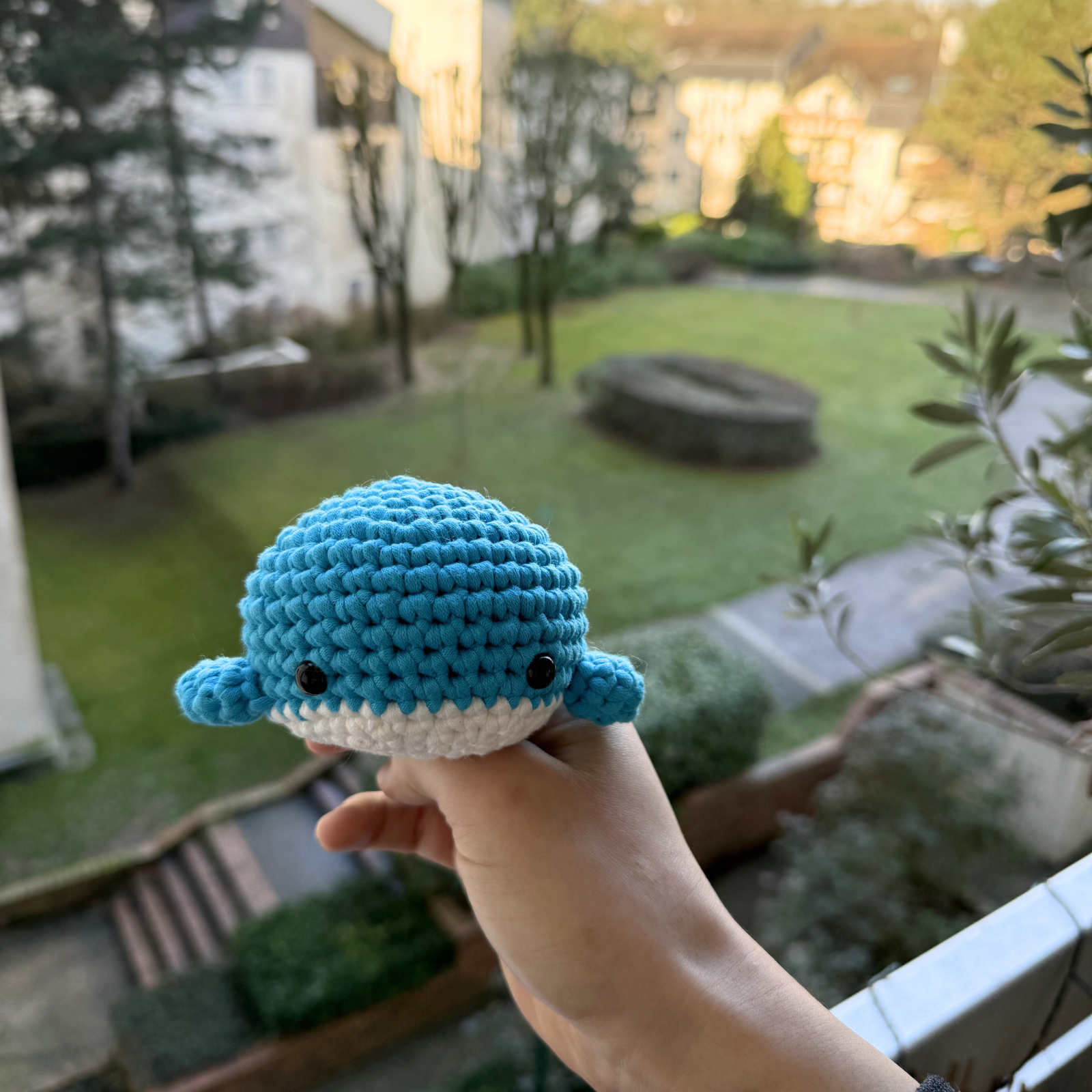 Hand holding a crocheted blue whale toy with a garden view in the background.