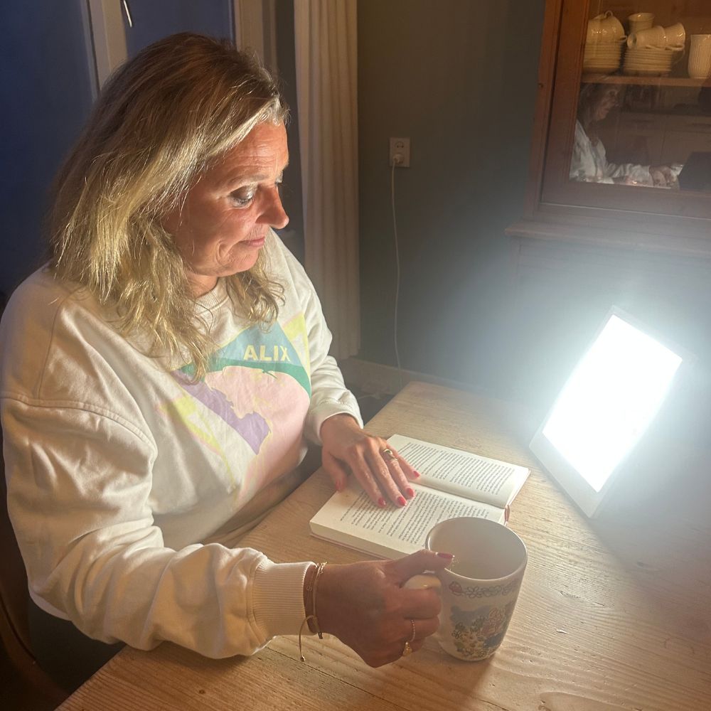 Woman reading a book at a table with a bright light and a cup.