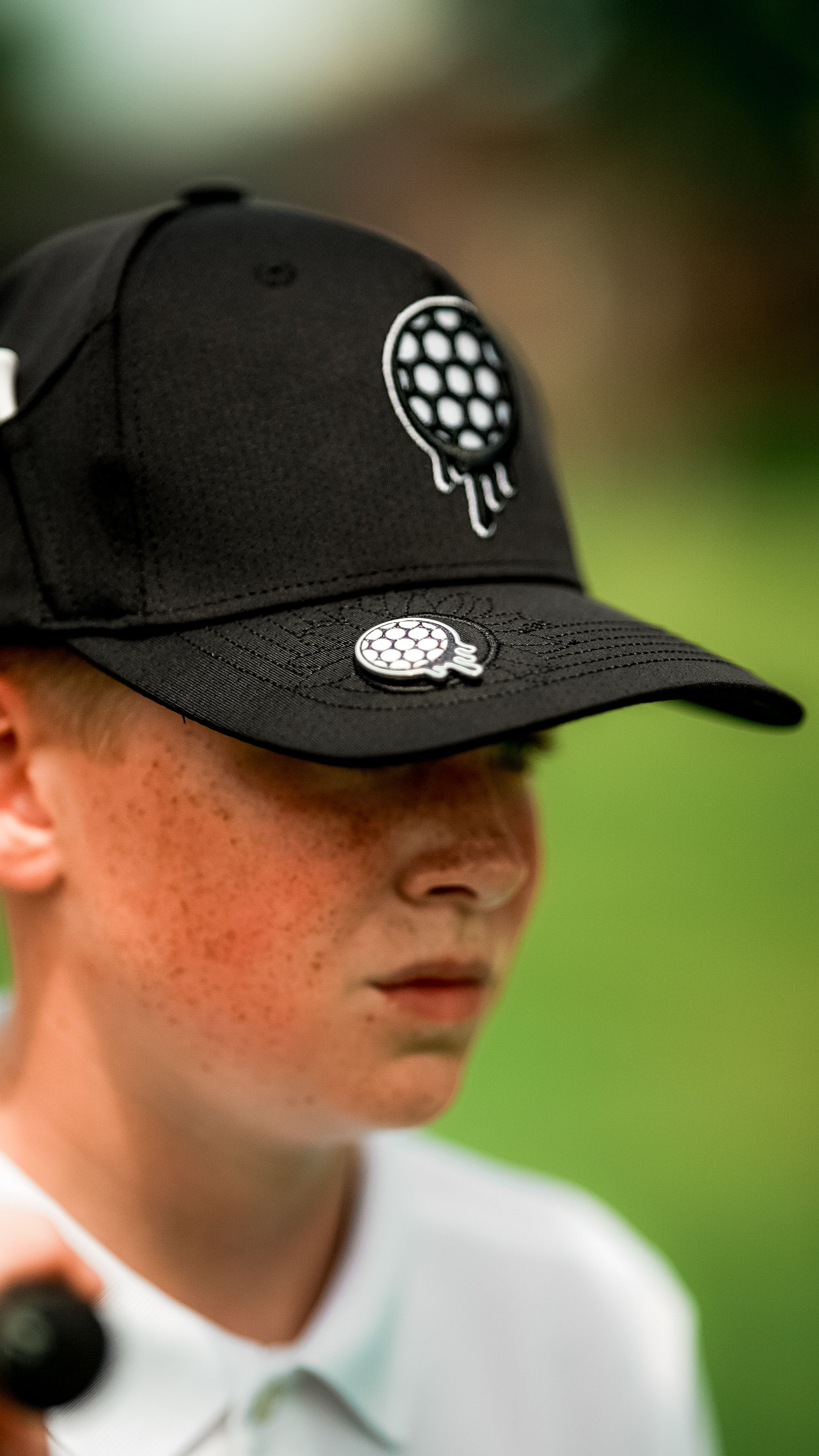 Person wearing a black cap with a golf logo, background is blurred greenery.
